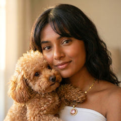 A woman with dark hair smiling while holding a small curly-haired brown dog, wearing a white strapless top and a gold necklace with a pink and white pendant.