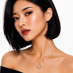 A close-up of a woman with short black hair wearing a delicate gold chain necklace with a round pendant featuring a pink gemstone surrounded by small clear stones, set against a plain white background.