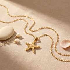 A gold starfish pendant necklace on a matching chain, styled on a beige linen fabric with a white stone, dried flowers, and a pink shell.