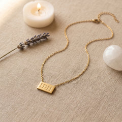 A gold chain necklace with a rectangular pendant engraved with the Roman numeral 'III' is displayed on a textured beige linen surface. The scene is styled with a lit white candle, a sprig of lavender, and a white stone.