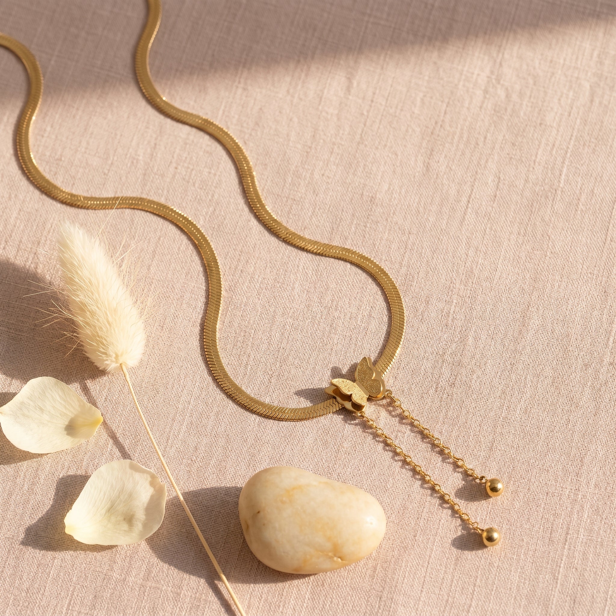 A gold snake chain necklace with a small leaf charm and dangling chain ends, styled on a beige linen surface with dried bunny tail grass, petals, and a smooth stone.