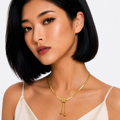 A close-up of a woman with a short black bob hairstyle wearing a delicate gold chain necklace with a bow-shaped centerpiece and dangling bead drops, paired with a cream camisole top against a light gray background.