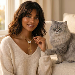 A woman wearing a gold necklace with a white butterfly and dangling key charms sits on a beige sofa next to a fluffy gray cat.