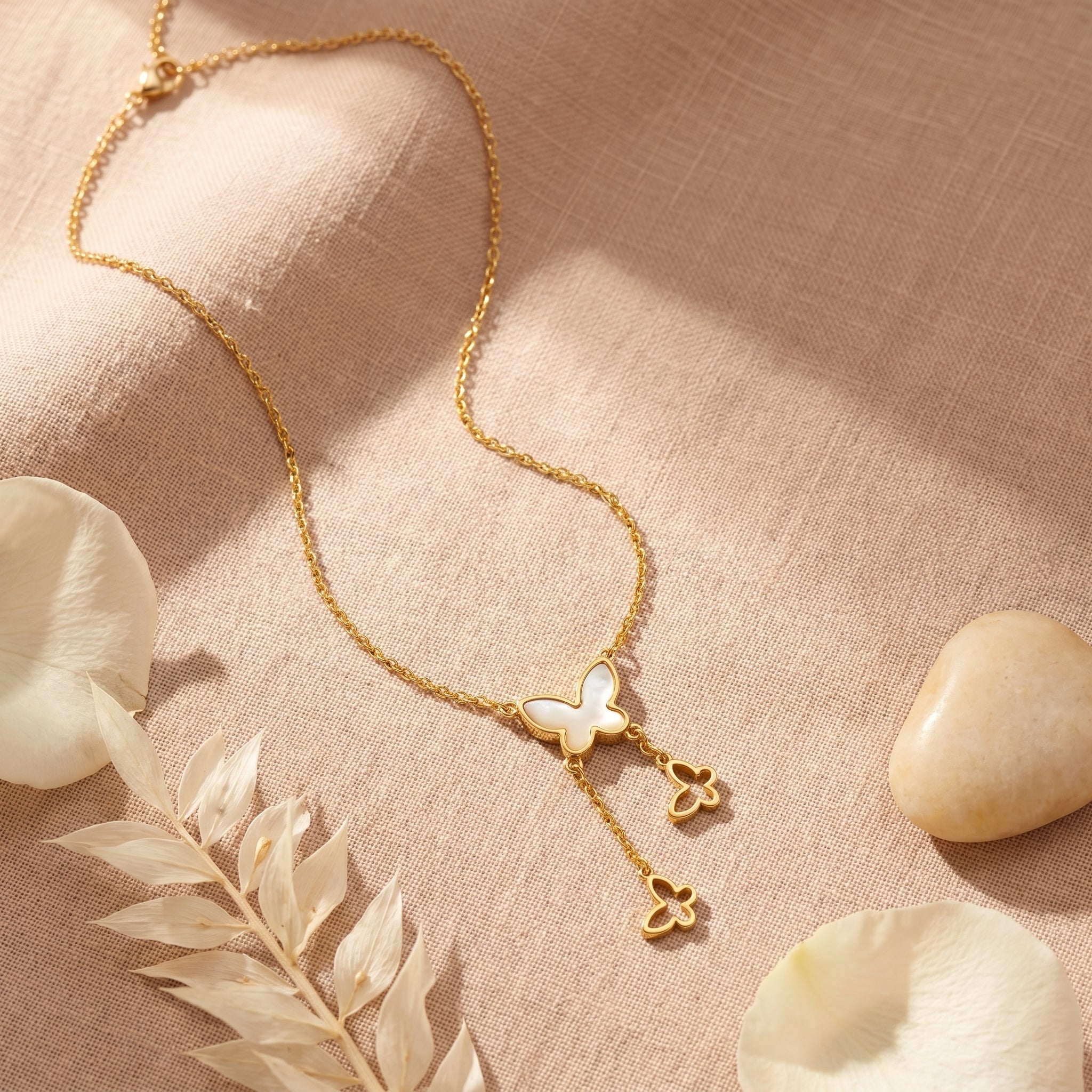 A delicate gold necklace with a butterfly and two small flower charms, displayed on a textured beige linen fabric with dried leaves and stones.
