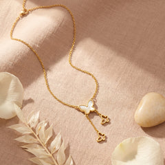 A delicate gold necklace with a butterfly and two small flower charms, displayed on a textured beige linen fabric with dried leaves and stones.