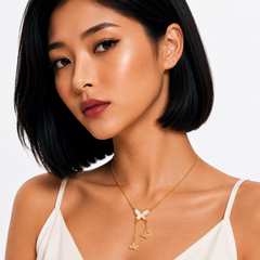 A close-up of a woman with short black hair wearing a delicate gold necklace with a white butterfly pendant and dangling charms, paired with a white camisole against a light gray background.
