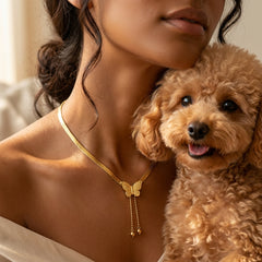 A close-up lifestyle shot of a woman wearing a delicate gold butterfly necklace, with a small curly-haired brown dog resting against her shoulder.