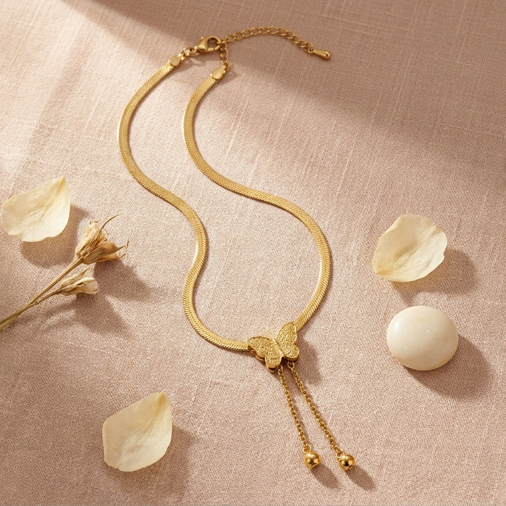 A gold snake chain necklace with a butterfly pendant and dangling chain ends, styled on a textured beige fabric with scattered flower petals and a white stone.