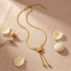 A gold snake chain necklace with a butterfly pendant and dangling chain ends, styled on a textured beige fabric with scattered flower petals and a white stone.