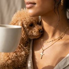 A close-up lifestyle shot of a woman wearing gold cross necklaces, with a small curly-haired brown dog resting against her shoulder and a white mug partially visible on the left.