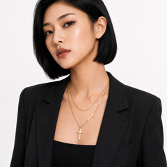 A woman with a short black bob hairstyle wears a layered gold necklace set featuring a larger cross pendant and a smaller cross charm, styled with a black blazer against a plain white background.