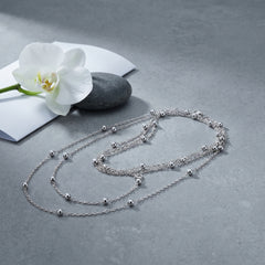 A delicate silver multi-strand necklace with small bead accents is arranged on a textured gray surface next to a white orchid flower and a dark stone.