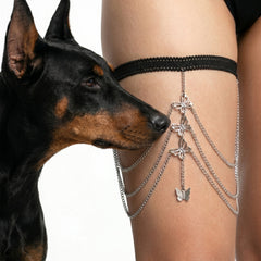 A black lace thigh strap with dangling silver chains and butterfly charms worn on a person's thigh, with a black-and-tan dog looking at the product against a white background.
