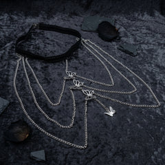 A gothic-style layered necklace featuring a black braided choker collar and multiple silver-tone chains with butterfly and bird charms, displayed on a dark textured background with scattered slate chips.