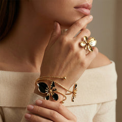 A close-up lifestyle shot of a model wearing gold jewelry, featuring a gold ring shaped like a flower and a gold cuff bracelet with black enamel butterfly and dragonfly details.