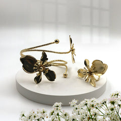Gold-tone flower-shaped cuff bracelet and matching flower-shaped ring displayed on a round white pedestal, with small white flowers in the foreground and a softly lit neutral background.