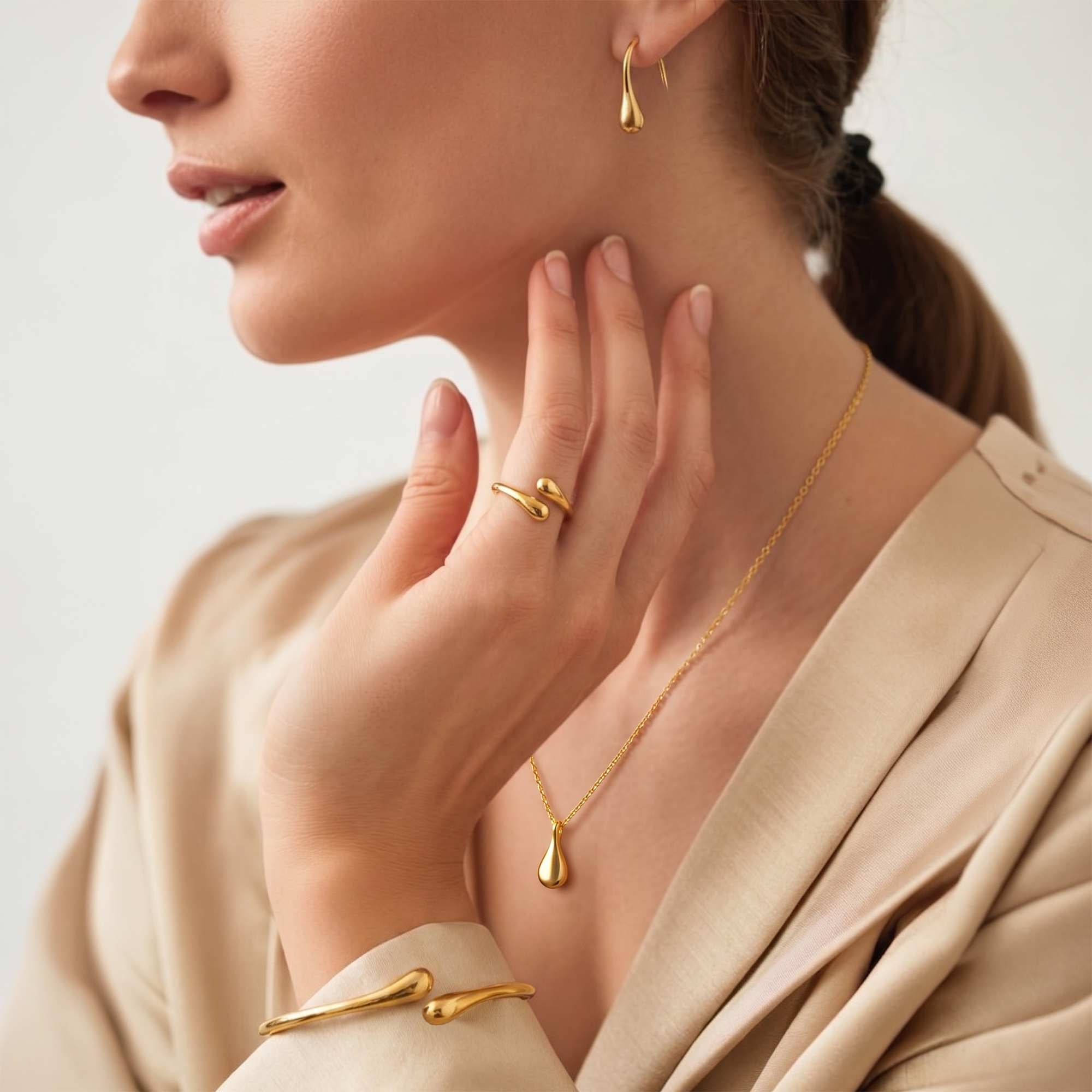 A close-up lifestyle shot of a woman wearing elegant gold jewelry, including a teardrop pendant necklace, a matching teardrop hoop earring, and a gold ring and bracelet with a twisted branch-like design.