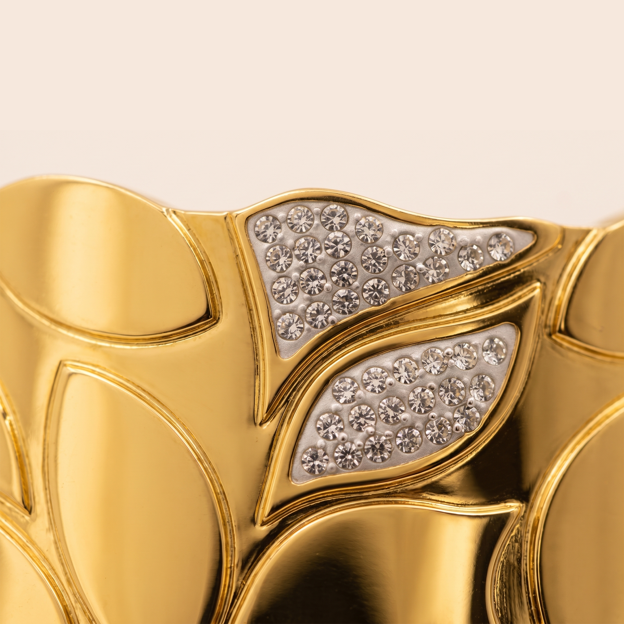 Close-up of a gold leaf-patterned cuff bracelet with two leaf-shaped sections set with clear pavé gemstones on a warm off-white background.
