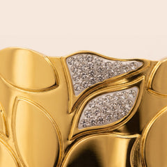 Close-up of a gold leaf-patterned cuff bracelet with two triangular sections set with clear pavé gemstones on a light background.