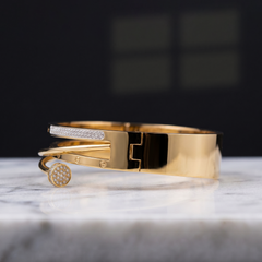 A polished gold-tone cuff bracelet with a pavé-set rectangular bar and a round pavé-set end, displayed on a white marble surface against a dark blurred background.