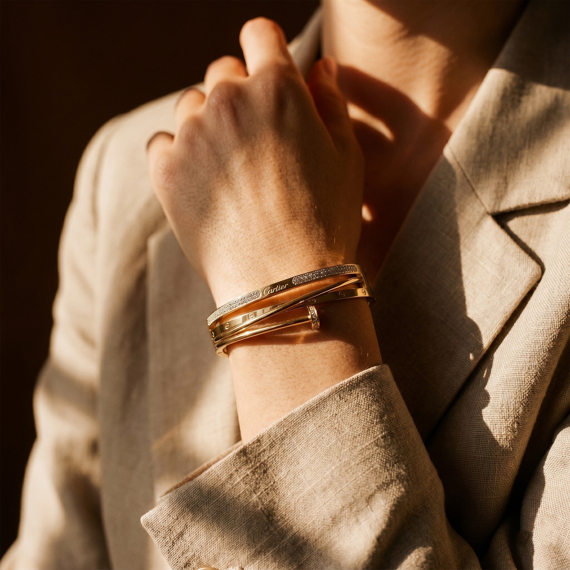 A close-up of a person wearing a stack of three gold bangles, including one with a textured pattern, set against a beige linen blazer with warm, dramatic sunlight.
