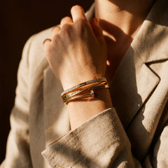 A close-up of a person wearing a stack of three gold bangles, including one with a textured pattern, set against a beige linen blazer with warm, dramatic sunlight.