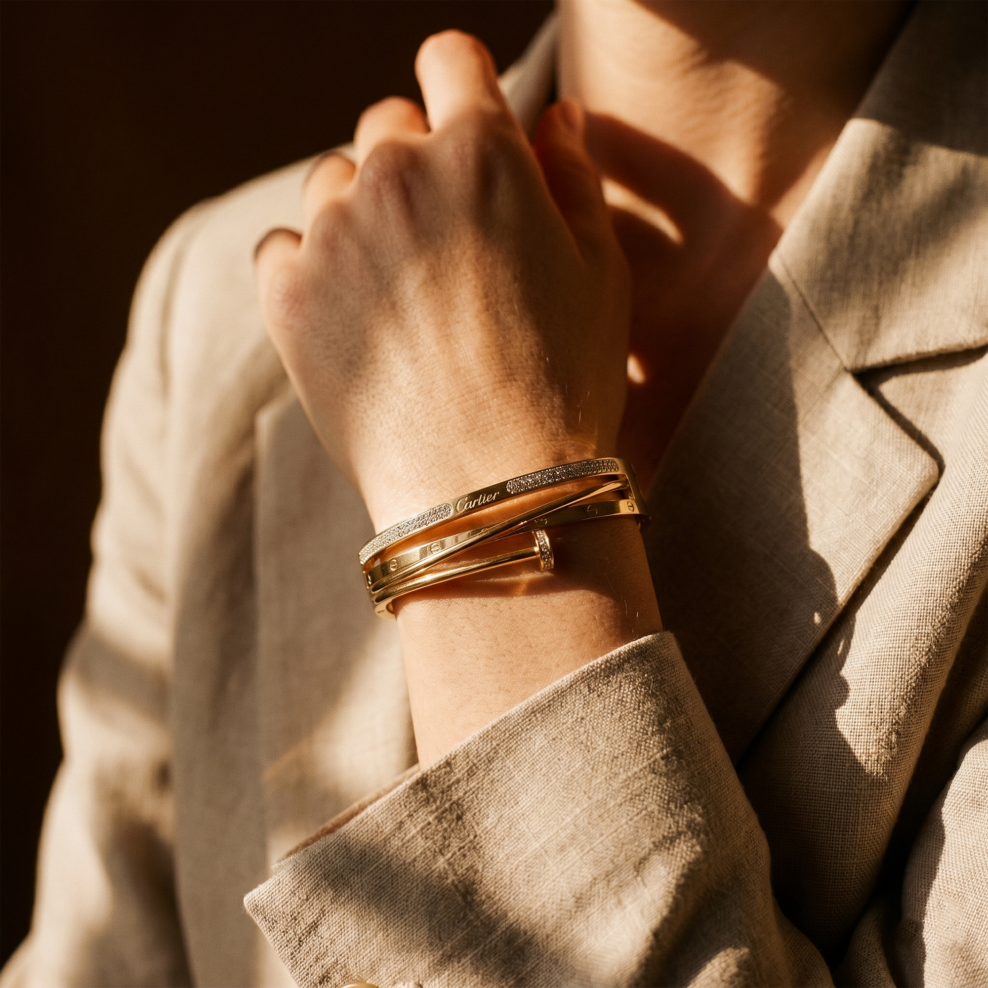 A close-up of a person wearing stacked gold bangles on one wrist, with warm sunlight casting shadows across their hand and beige blazer.