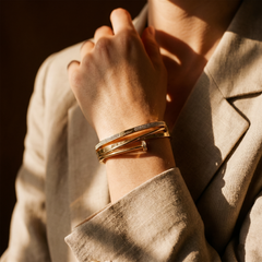 A close-up of a person wearing stacked gold bangles on one wrist, with warm sunlight casting shadows across their hand and beige blazer.
