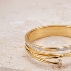 A gold bangle bracelet with diamond accents and a visible Cartier logo, photographed on a light beige stone surface.