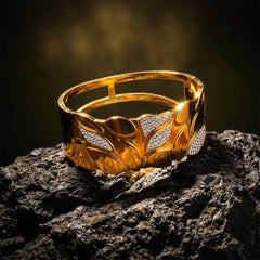 A wide gold cuff bracelet with a leaf pattern and diamond-set accents, displayed on a dark textured rock against a dark, moody background.