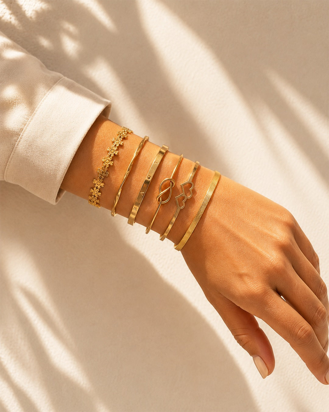 A close-up of a person's wrist and hand wearing five stacked gold bracelets against a warm cream background with soft leaf shadows.