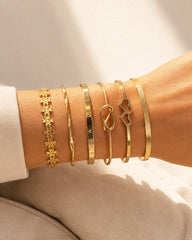 A close-up of a person's wrist wearing six gold-toned bracelets, including floral, bangle, engraved, infinity, and cloud-shaped designs, styled with a white sleeve and beige fabric background.