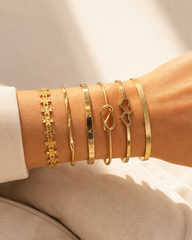 A close-up of a person's wrist wearing six delicate gold bangles, including floral, engraved, and openwork designs, styled with a white sleeve and beige fabric background.