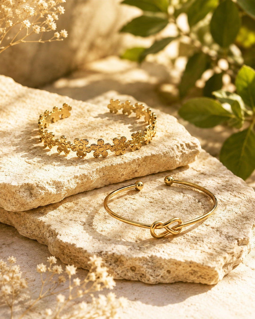 Two gold-toned bracelets displayed on textured beige stones with dried flowers and green leaves in a warm, sunlit setting. One bracelet is a flower-patterned cuff, and the other is a slim bangle with an infinity knot detail.
