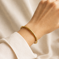 A gold bamboo-style bangle bracelet worn on a person's wrist, with a white long-sleeve shirt cuff visible against a soft beige fabric background.