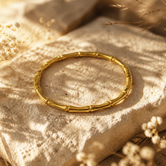A gold-toned bangle bracelet with a bamboo-inspired segmented design, displayed on a textured beige stone surrounded by dried white flowers under warm sunlight.