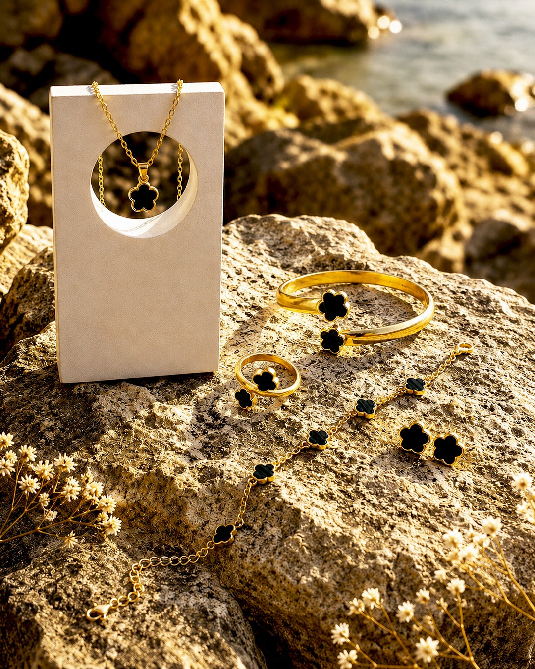 Gold jewelry set with black clover-shaped stones, including a necklace, bracelet, ring, and earrings, displayed on a rock outdoors next to a white jewelry box.