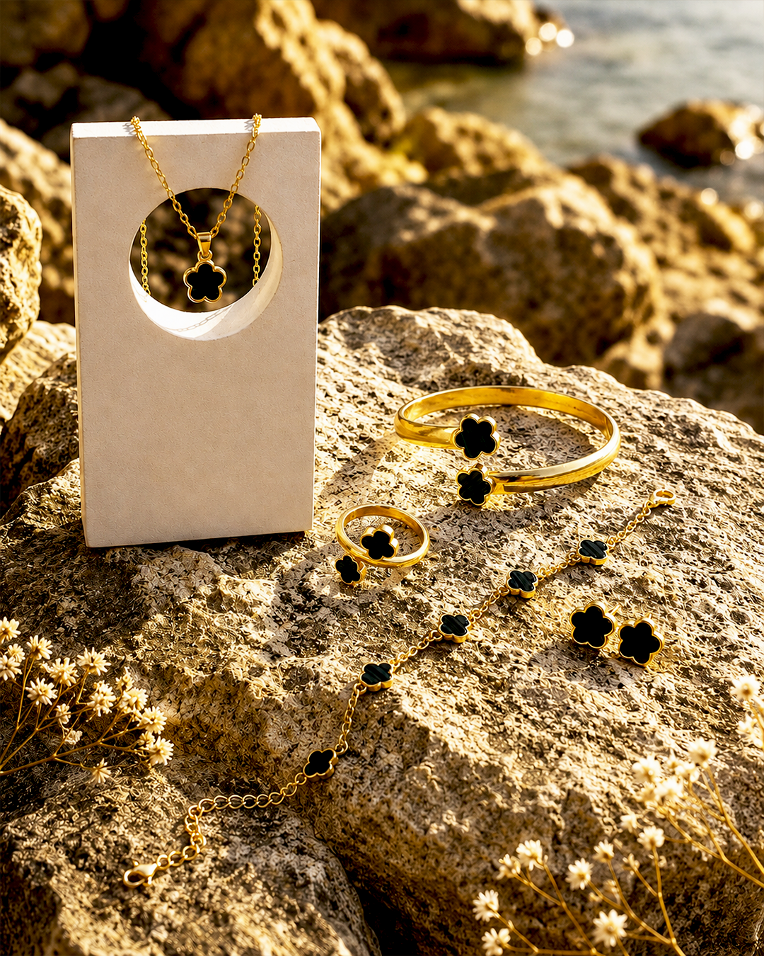Gold jewelry set with black flower-shaped accents, including a necklace, bracelet, ring, and earrings, displayed on a rock next to a white jewelry box.