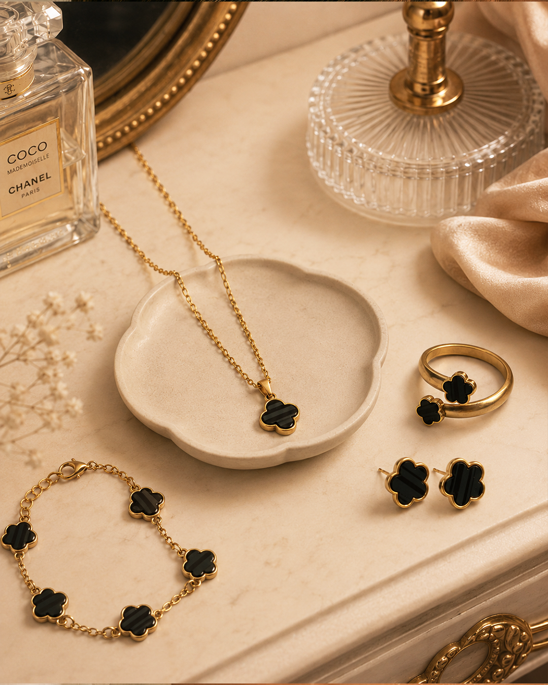 Gold jewelry set with black flower-shaped accents, including a necklace, bracelet, ring, and earrings, arranged on a beige surface with a small dish, perfume bottle, and glass decor.