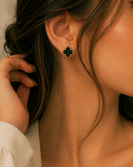 A close-up of a woman's ear wearing a small black clover-shaped stud earring with a thin gold-tone outline.