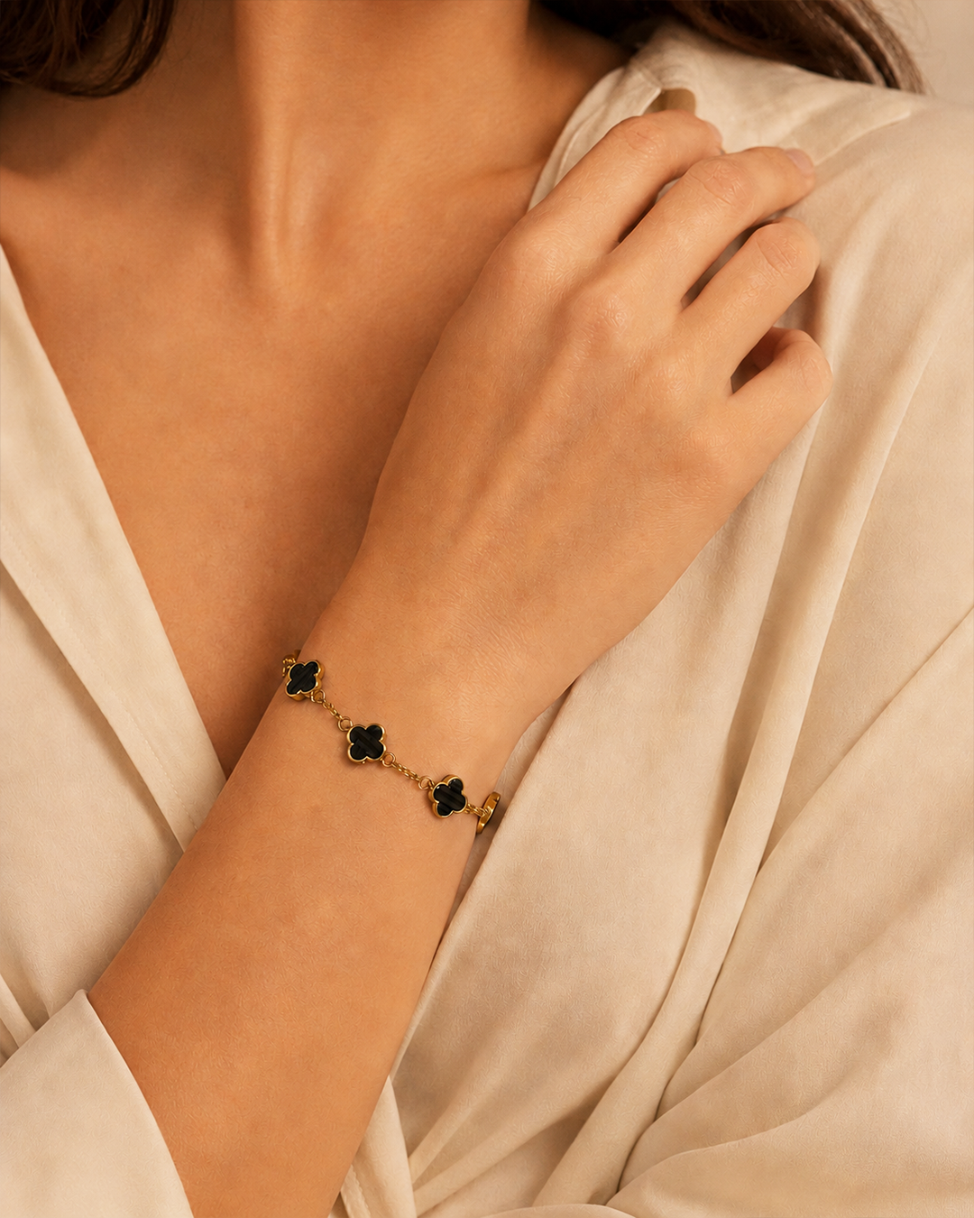 A close-up of a woman's hand and wrist wearing a delicate gold chain bracelet with four black clover-shaped charms, styled with a cream-colored satin blouse.