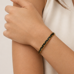 A close-up of a person's wrist wearing a thin black bangle bracelet with gold detailing and a small gold clasp, paired with a white sleeveless top.