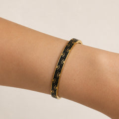 A gold bangle bracelet with a black enamel background and repeating gold monogram pattern, shown worn on a person's wrist against a light beige background.
