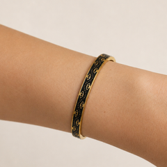 A gold bangle bracelet with a black enamel background and repeating gold monogram pattern, shown worn on a person's wrist against a light beige background.