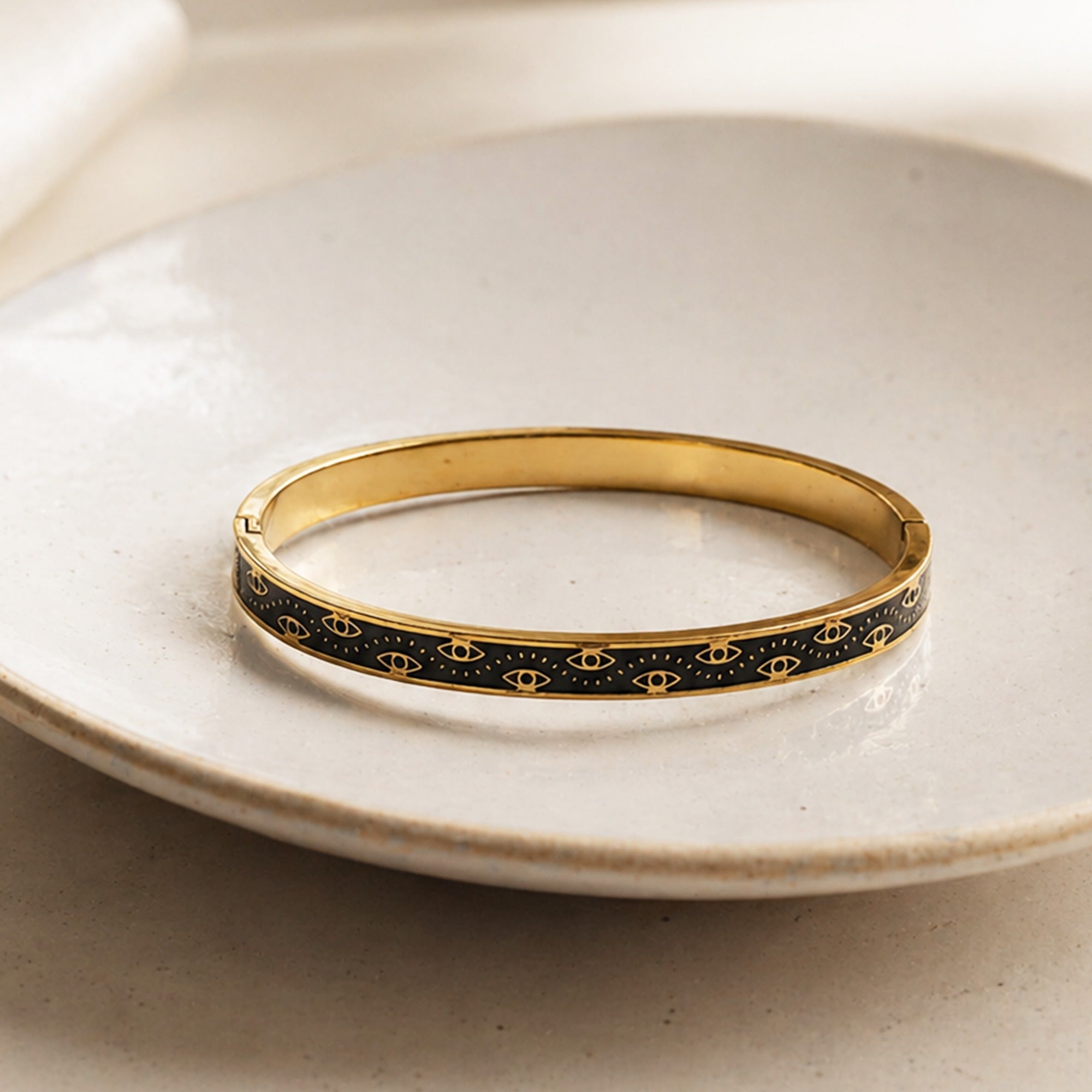 A gold bangle bracelet with a black enamel inlay featuring a repeating gold eye motif, resting on a light beige ceramic plate.