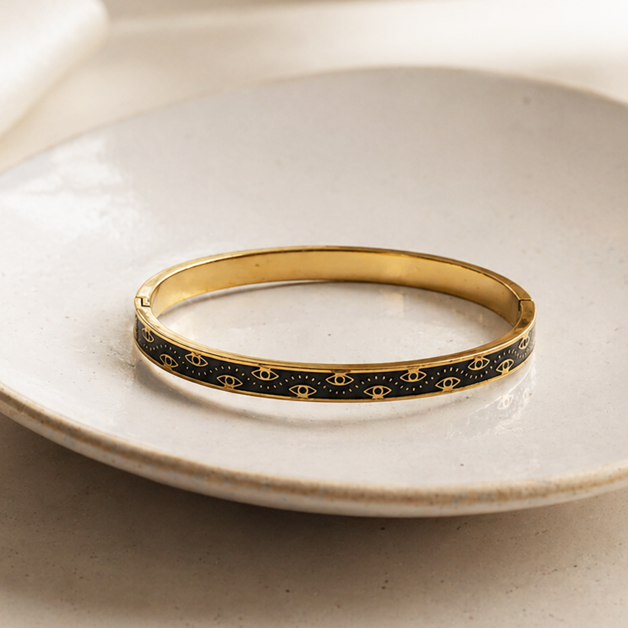 A gold bangle bracelet with a black enamel inlay featuring a repeating gold eye motif, resting on a textured beige ceramic plate.