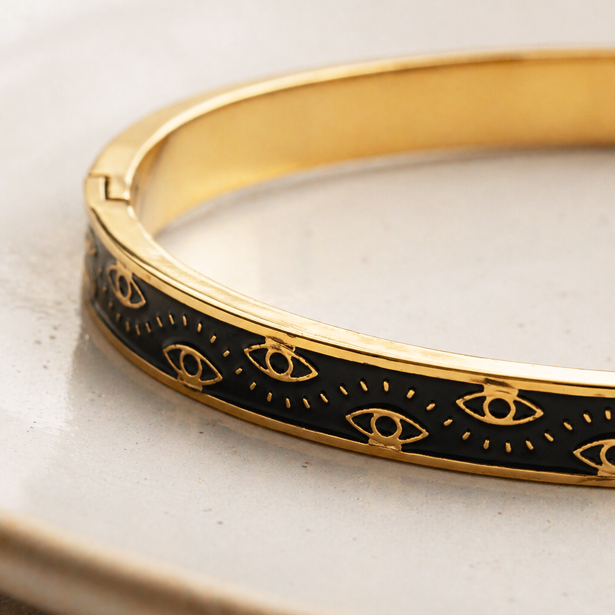 A gold-plated hinged bangle bracelet with a black enamel inlay featuring a repeating gold Eye of Providence pattern, resting on a light beige ceramic surface.