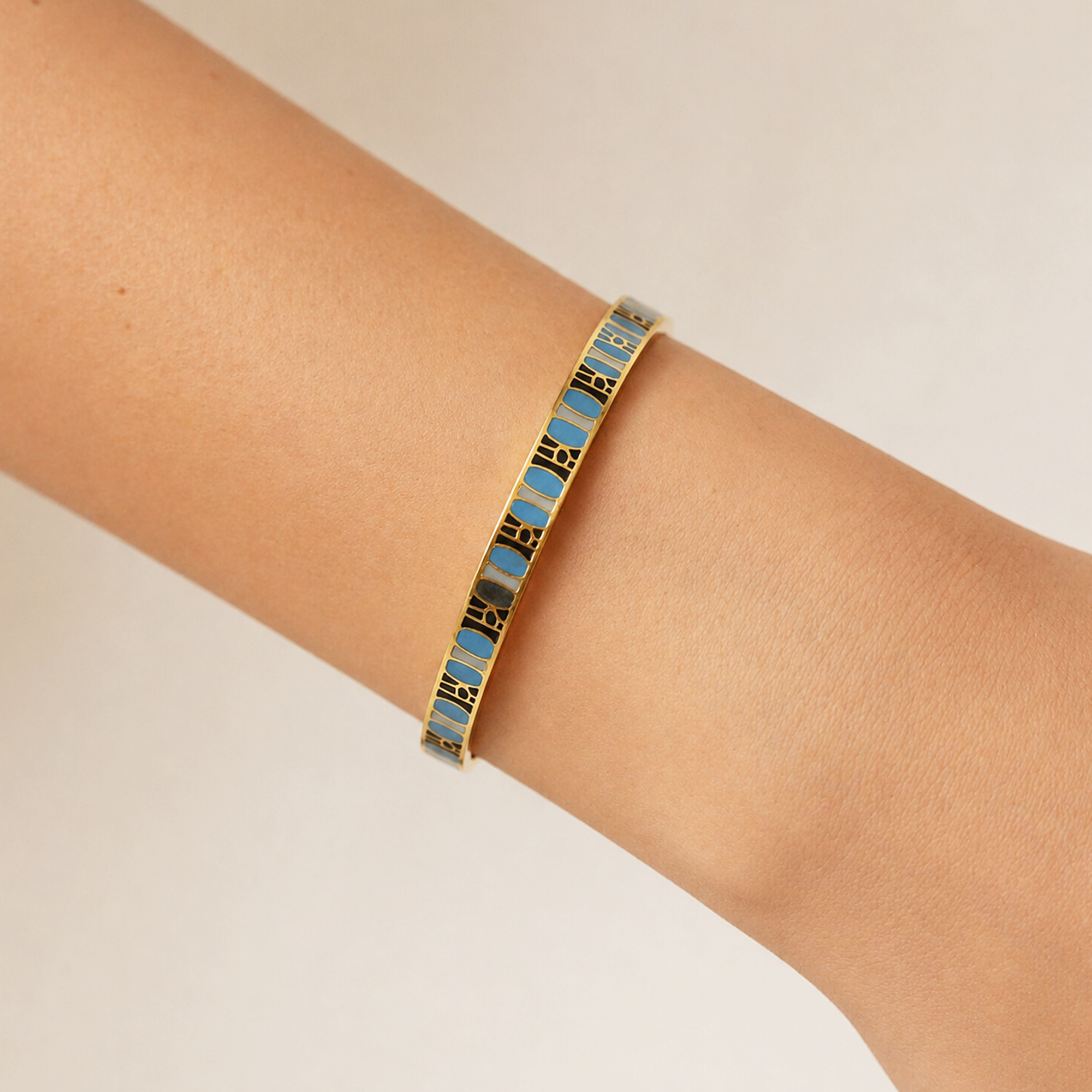 A gold-plated bangle bracelet with alternating light blue and black enamel segments, shown worn on a person's wrist against a plain light beige background.