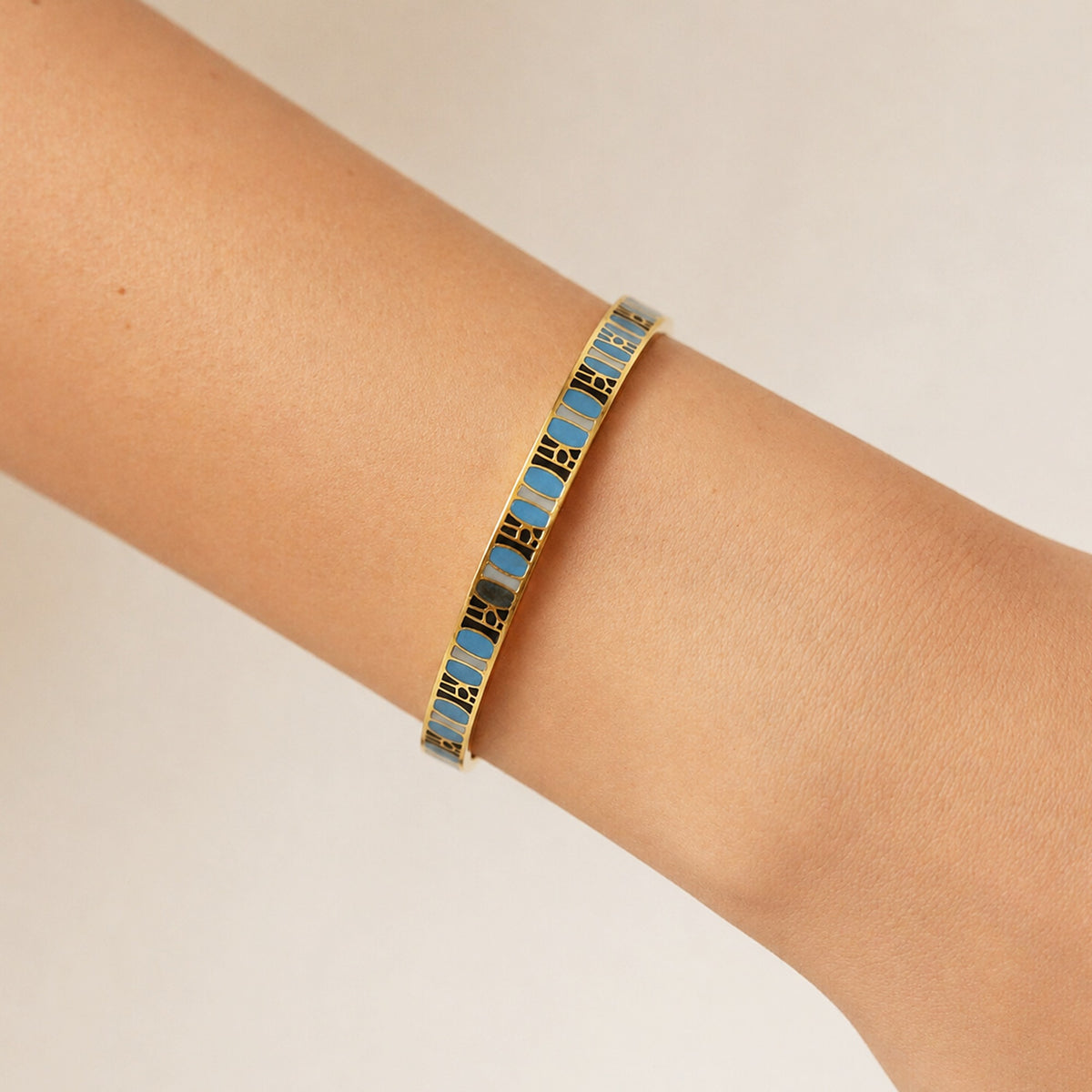 A gold-plated bangle bracelet with alternating light blue and black enamel segments, shown worn on a person's wrist against a plain light beige background.