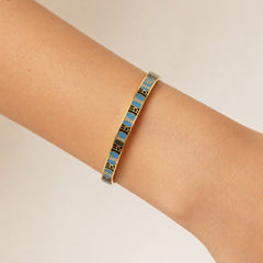 A gold-plated bangle bracelet with alternating light blue and black enamel segments, shown worn on a person's wrist against a plain light beige background.